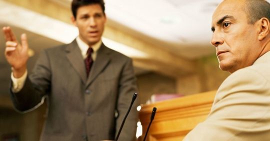Two men in a courtroom, one speaking while another sits at a wooden bench listening
