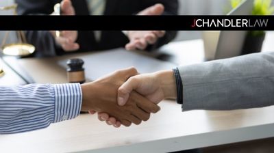 Two people in business attire shaking hands across a desk, with a blurred office background.