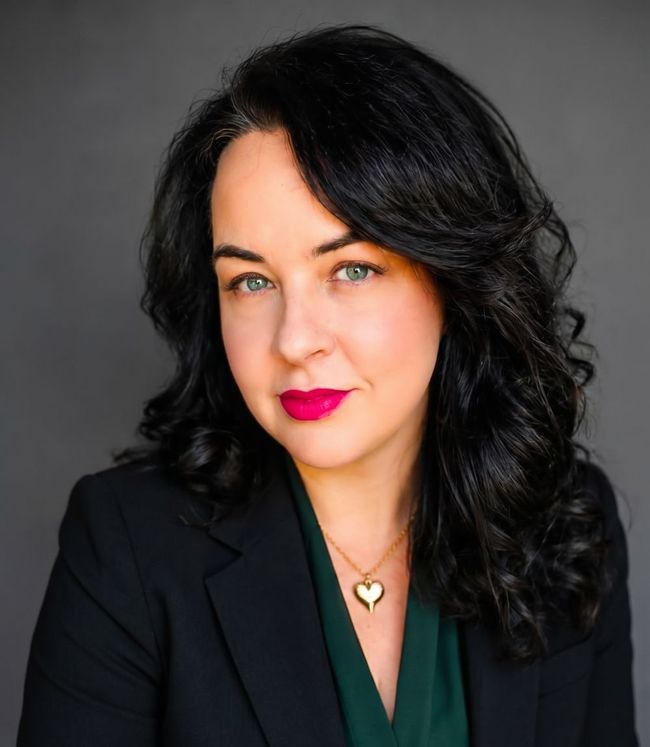 Portrait of a woman with black hair in a black blazer, wearing pink lipstick against a gray background