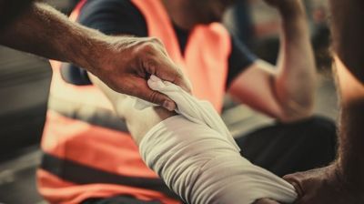 Hands wrapping a bandage around an injured arm during a first-aid event, with a worker in an orange safety vest in the background