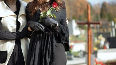 Two people in black dresses and gloves holding a red rose at an outdoor memorial.