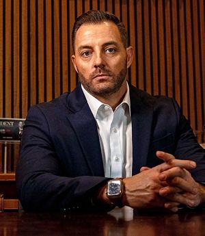 Man in navy suit seated at a table against a wood-paneled wall, looking directly at the camera.