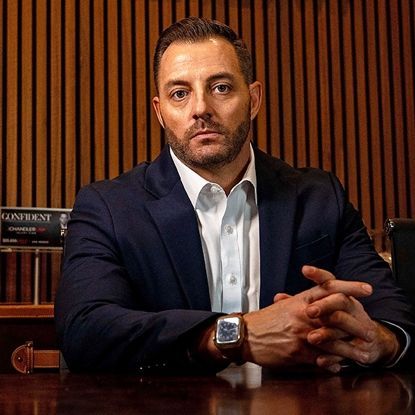 Man in a navy suit seated at a desk, hands clasped, against a wood-paneled wall