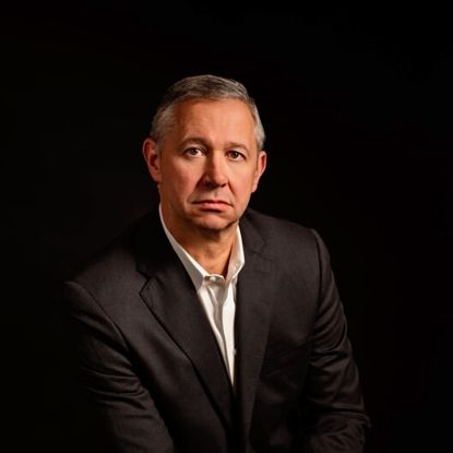 Studio portrait of a man in a dark suit against a black background, facing forward with a neutral expression