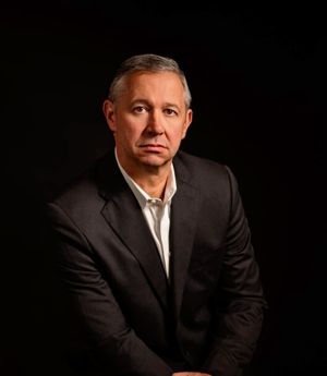 Studio portrait of a man in a dark suit against a black background, looking at the camera