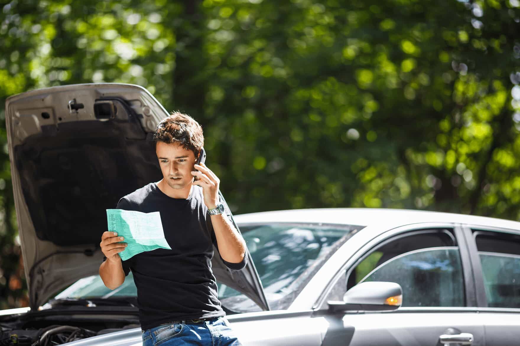 Man reading a paper beside a broken-down car with the hood open, holding a phone on a sunny roadside