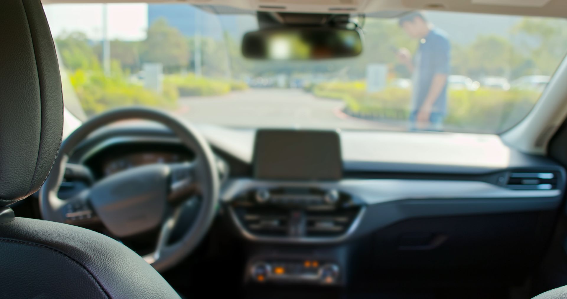 View from inside a car over the dashboard and steering wheel toward a person walking outside on a sunny street.