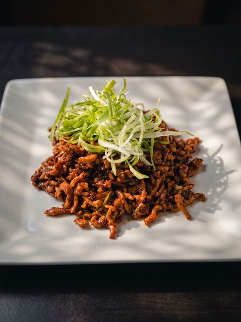 A square white plate with saucy minced meat topped with shredded green onions, in sunlight on a dark table