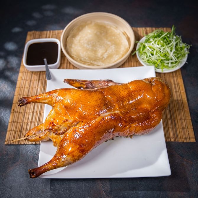Roasted whole chicken glazed golden brown on a plate with dipping sauce, soup, and greens on a bamboo mat.