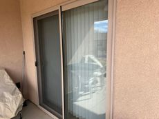 Sliding glass patio door with vertical blinds and a reflected car, next to a covered item on a stucco wall