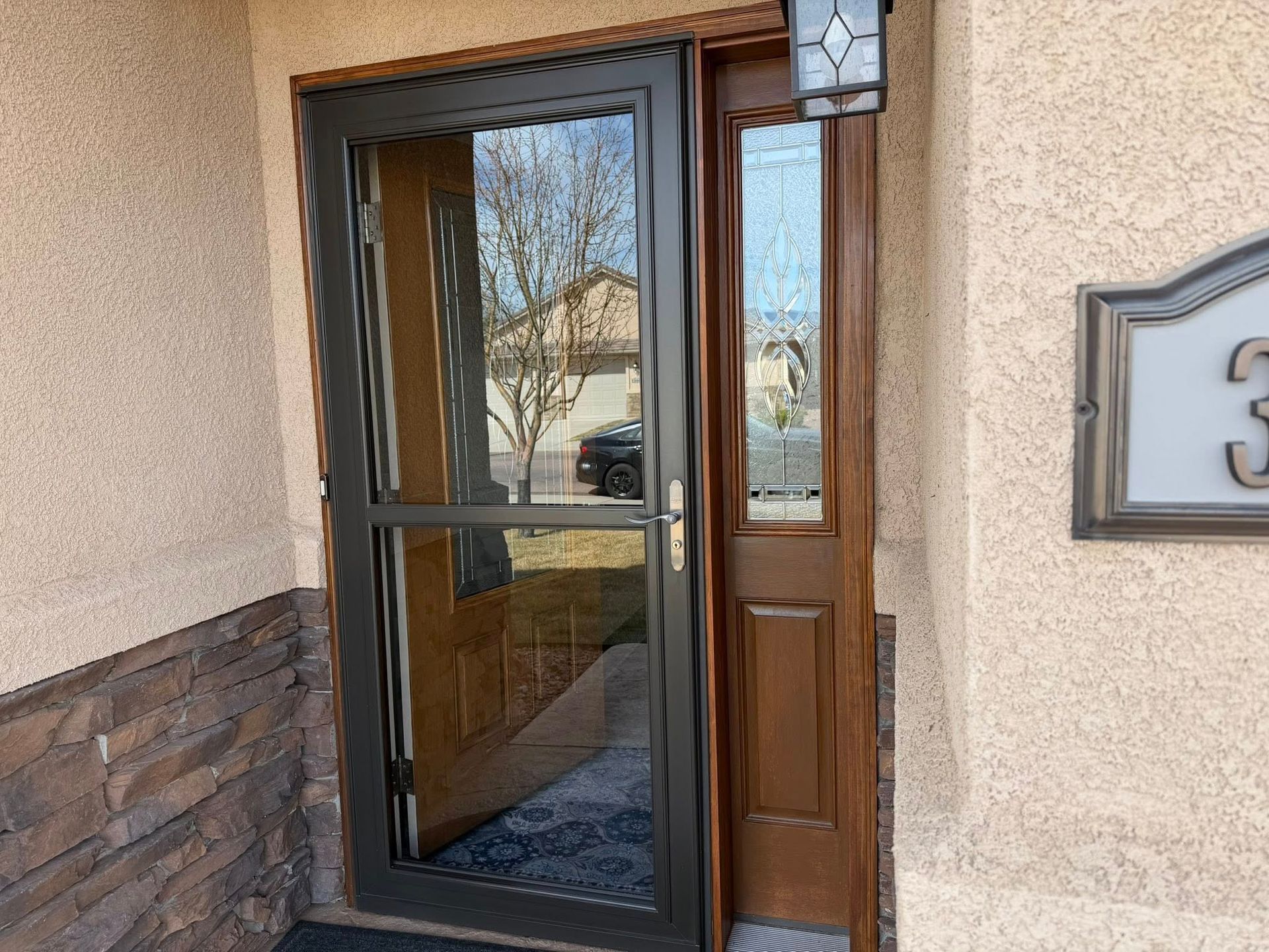 Front entrance with glass storm door, wooden door, and house number plaque beside a stucco wall.