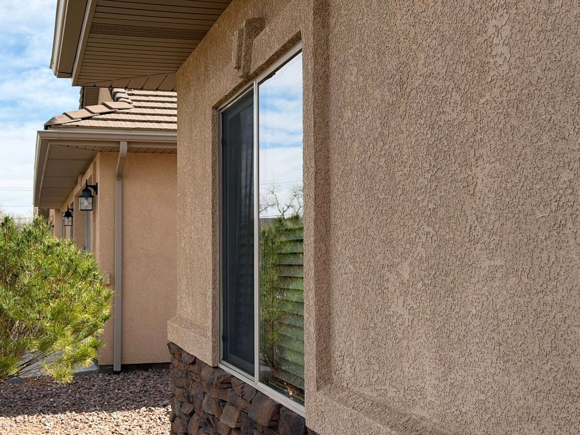 Side view of a tan stucco house with a window, roof eaves, and desert landscaping.