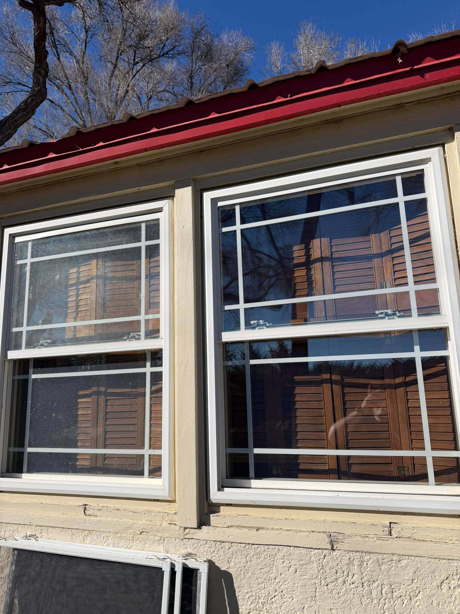 Two white-trimmed windows on a beige house under a red roof, with bare tree branches above.