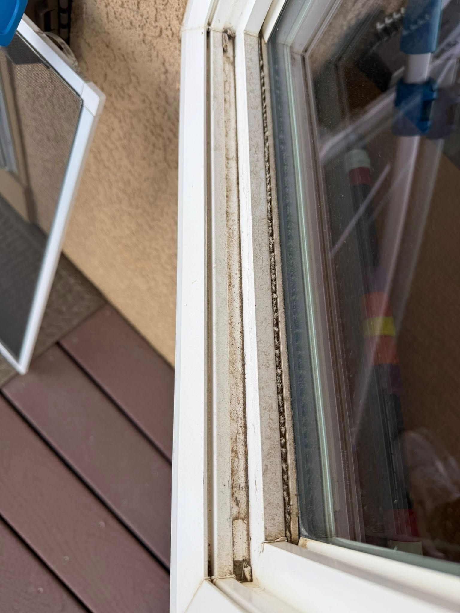 Close-up of a dirty white window frame beside brown siding and a deck, with a window reflection.