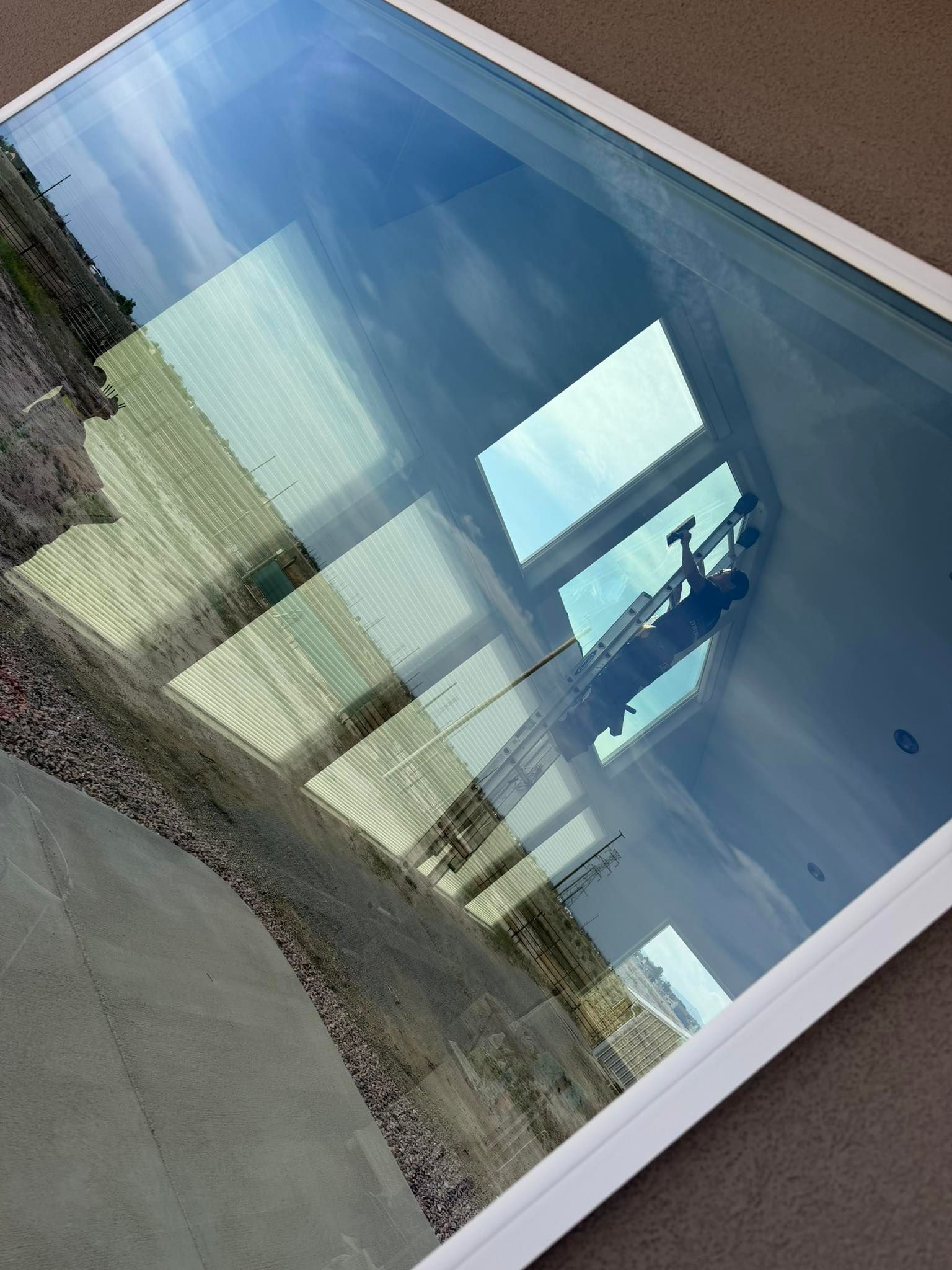 Tilted view of a glass window reflecting sky, clouds, and nearby buildings beside a concrete walkway.