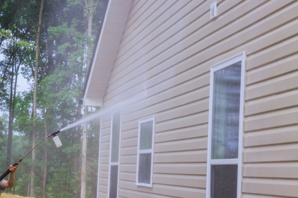 Person pressure-washing a beige house exterior beside trees and windows