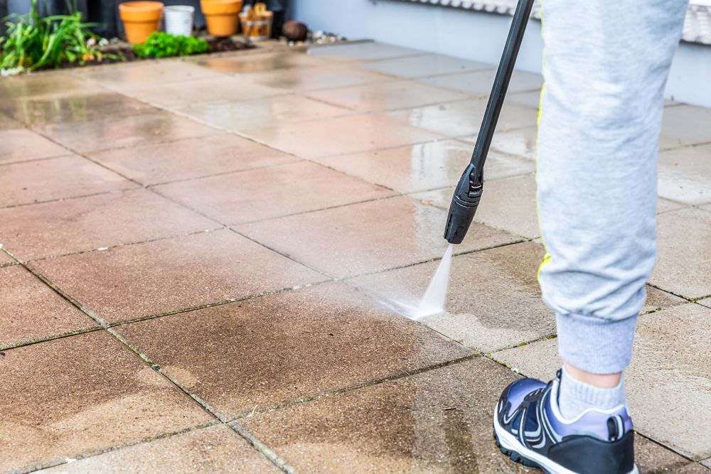 Person pressure-washing a wet patio with a spray nozzle, wearing gray sweatpants and blue sneakers