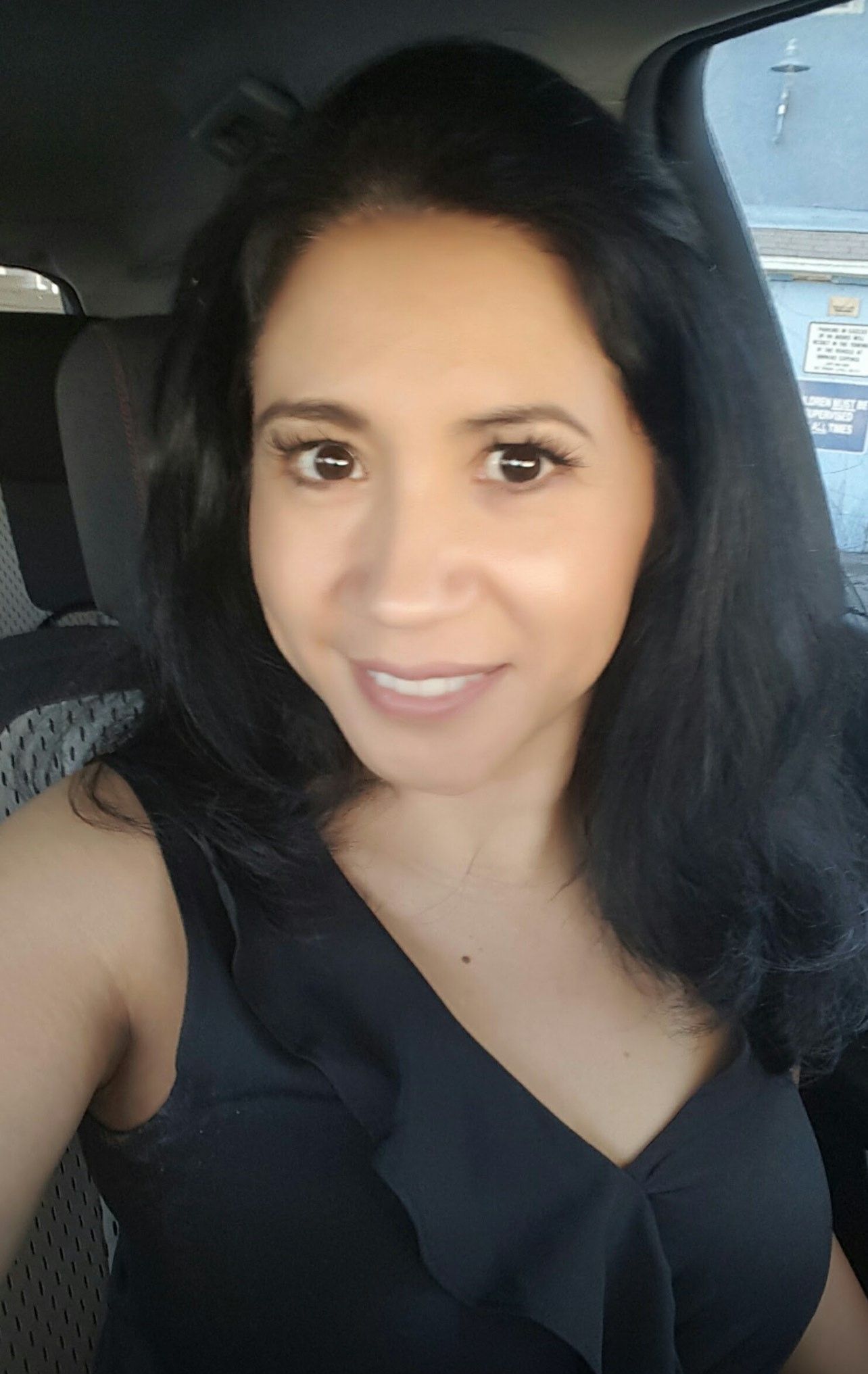 Woman smiling in a black top, sitting in a car.