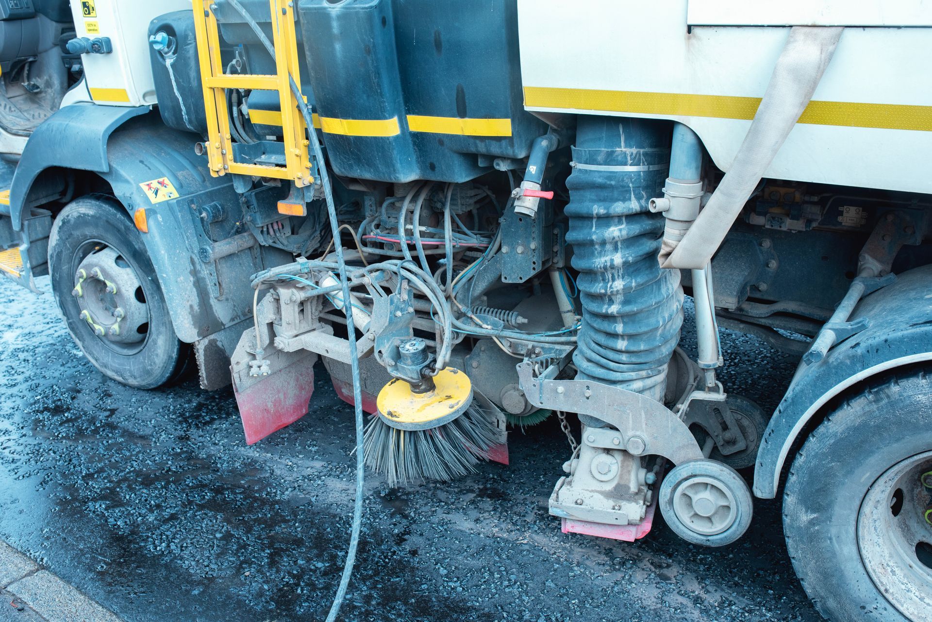 Close-up of a mechanical sweeper truck's rotating brush and vacuum hose on pavement.
