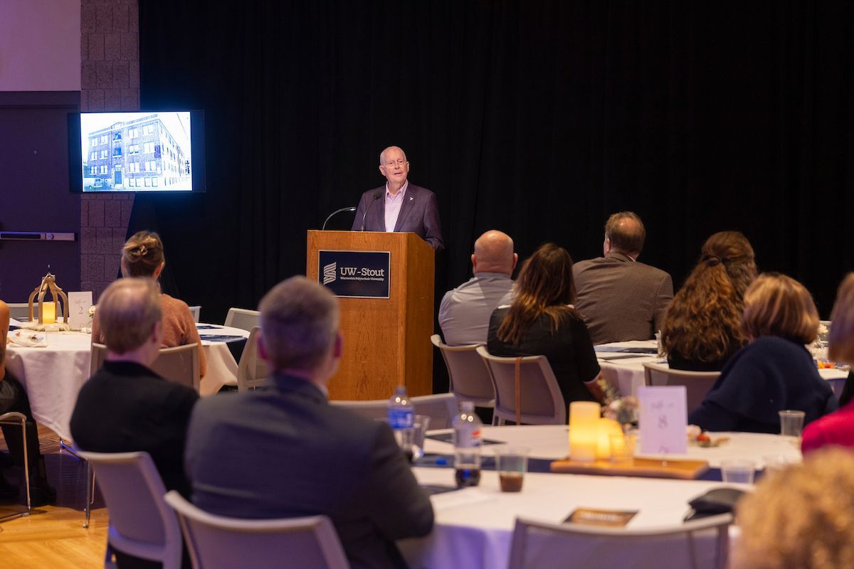 A speaker stands at a podium addressing an audience in a dim, banquet-style room with a screen showing a building photo.