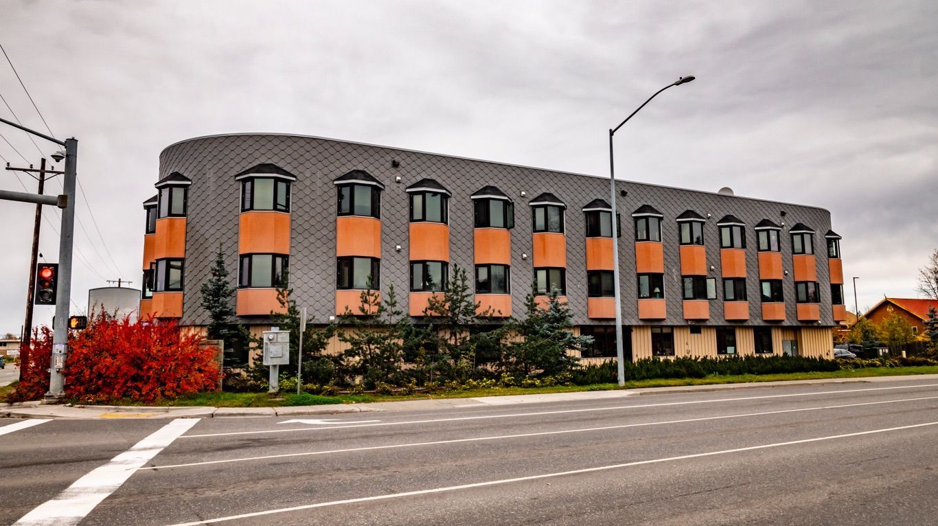 A multi-story commercial building with grey tile and orange accents stands at a street corner under a cloudy sky.