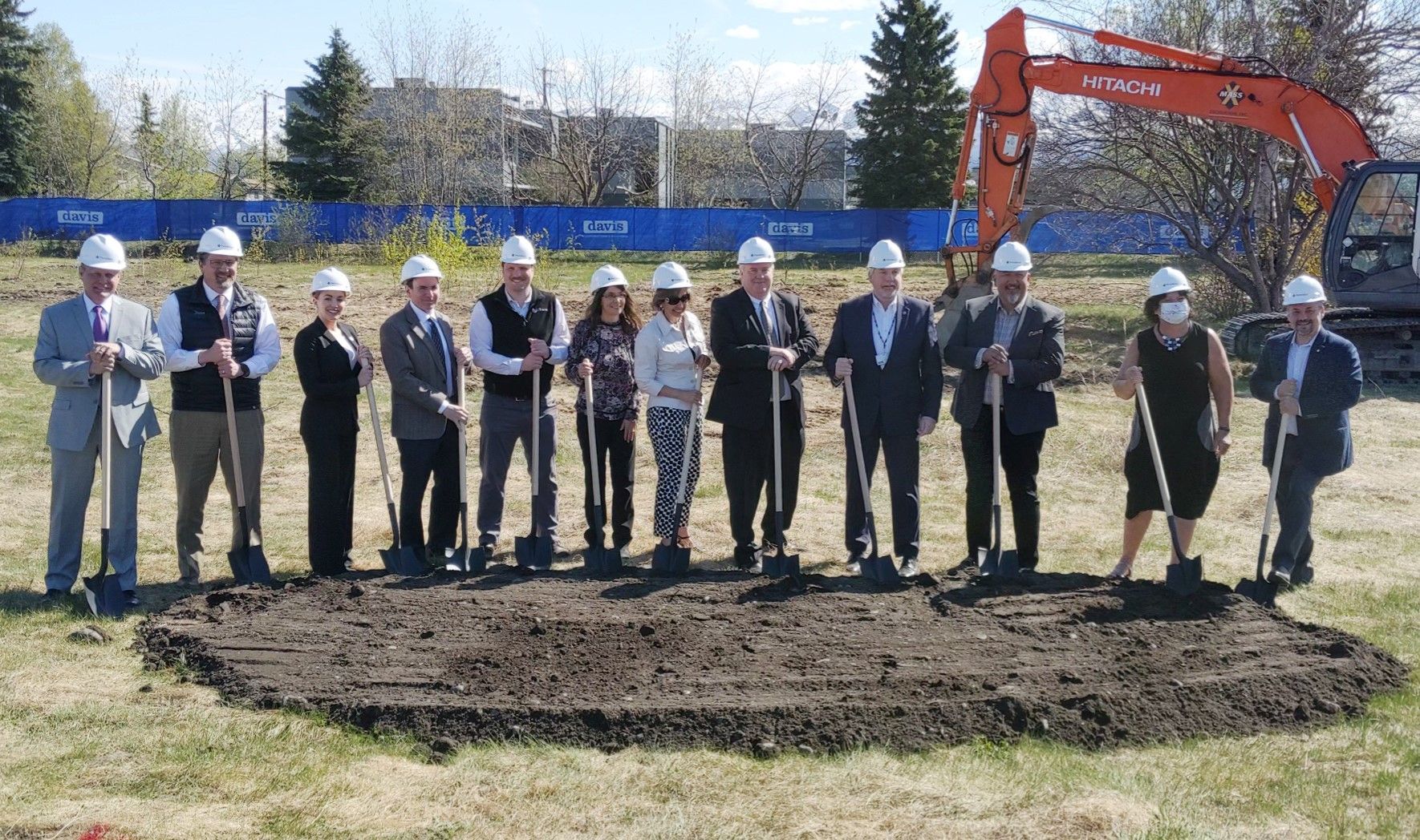 A group of people in hard hats stand in a row outdoors, holding shovels for a ceremonial ground-breaking at a construction site.