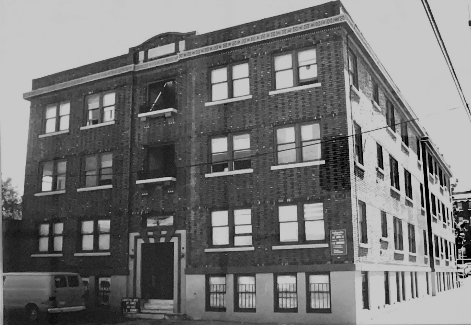 A low-angle, black-and-white view of a multi-story brick apartment building with a white van parked in the foreground.