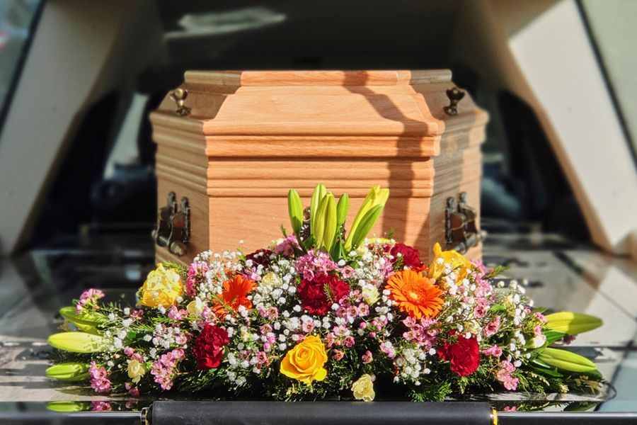 Wooden casket with colorful flowers arranged in front, seen at a funeral.