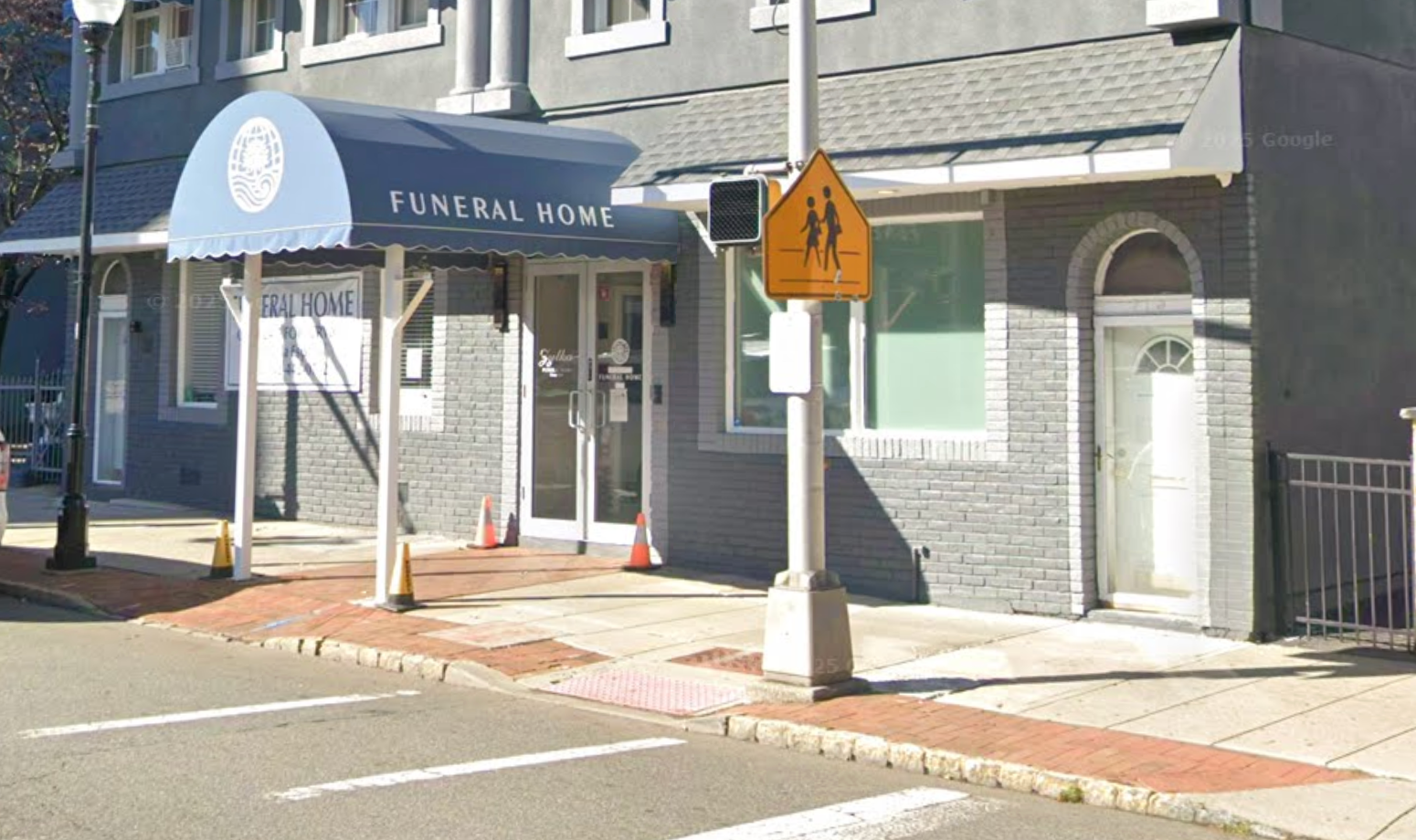 Funeral home with blue awning on a sunny street corner, beside a pedestrian crossing sign