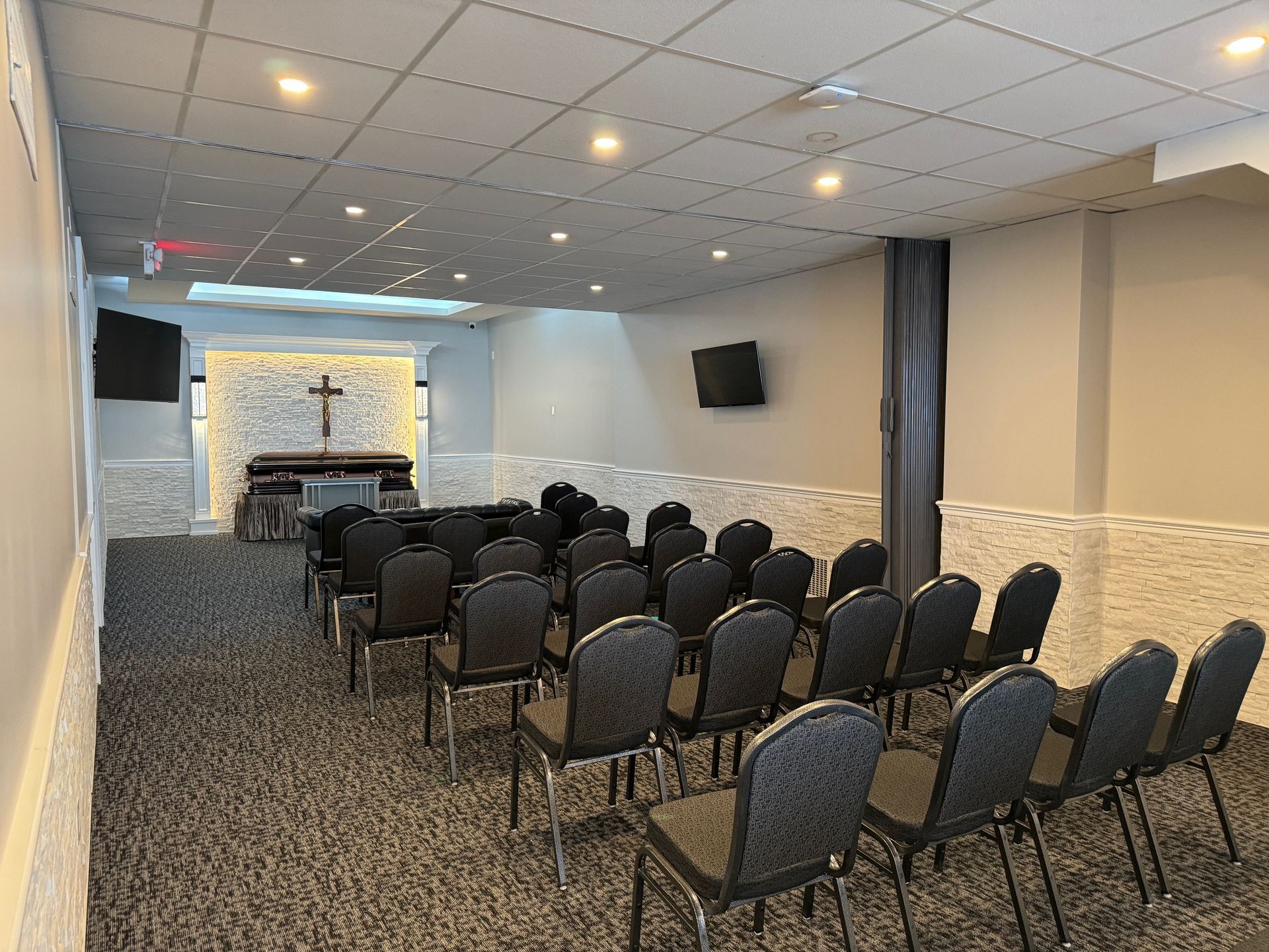 Small chapel with rows of gray chairs facing a lit altar and cross in a carpeted room