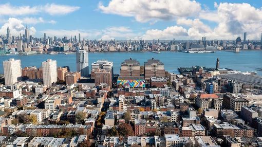 Aerial view of a city skyline across the river, with dense buildings and a bright blue sky with clouds.