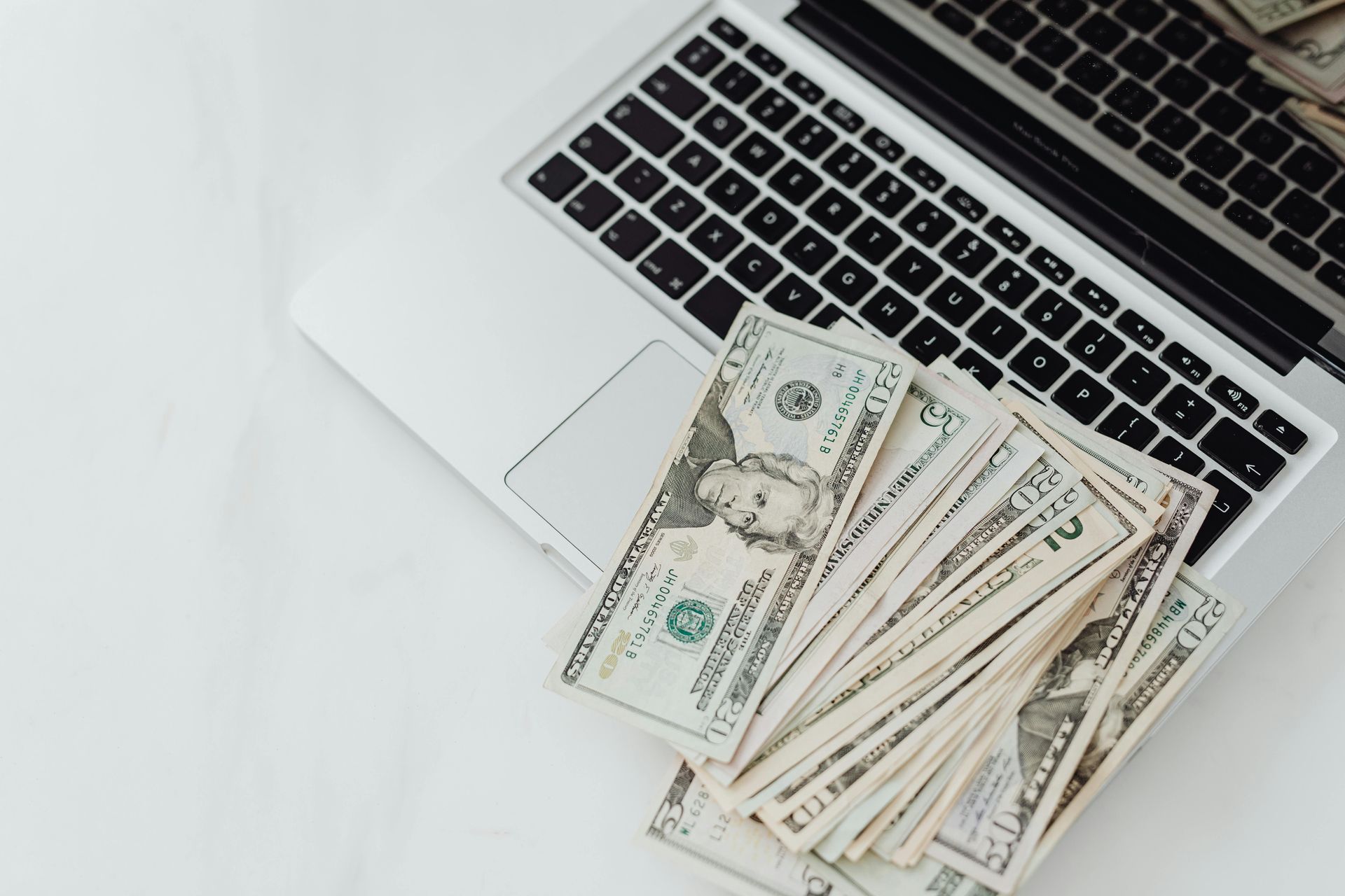 Cash laid on a laptop keyboard next to a white notebook on a desk