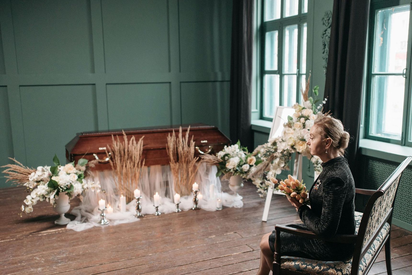 Woman seated in a teal room beside floral arrangements and a wooden coffin