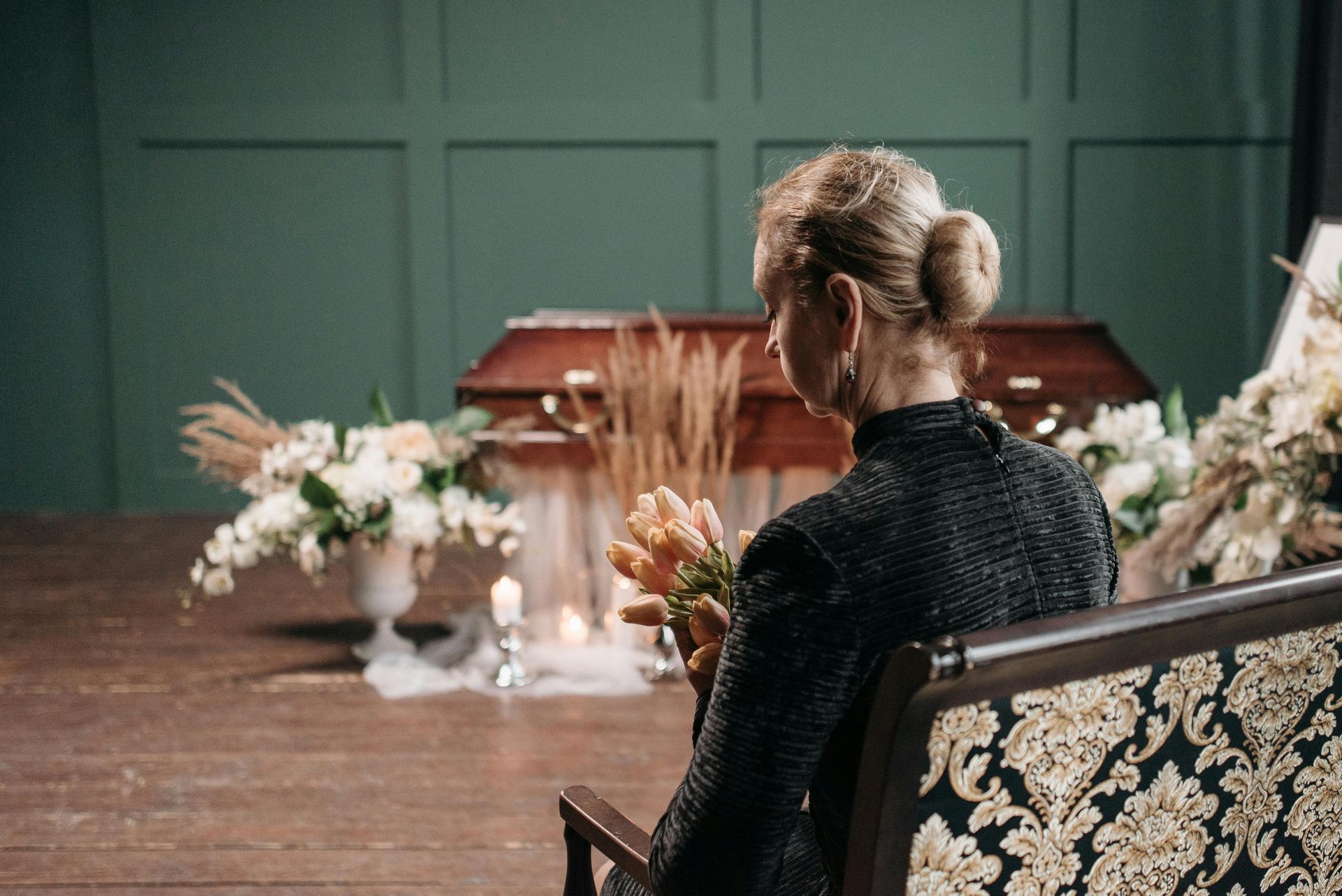 Woman seated at a long table in a floral room, facing a closed casket with flowers nearby