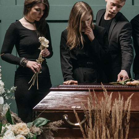 Mourners in black stand beside a casket with flowers at a funeral.