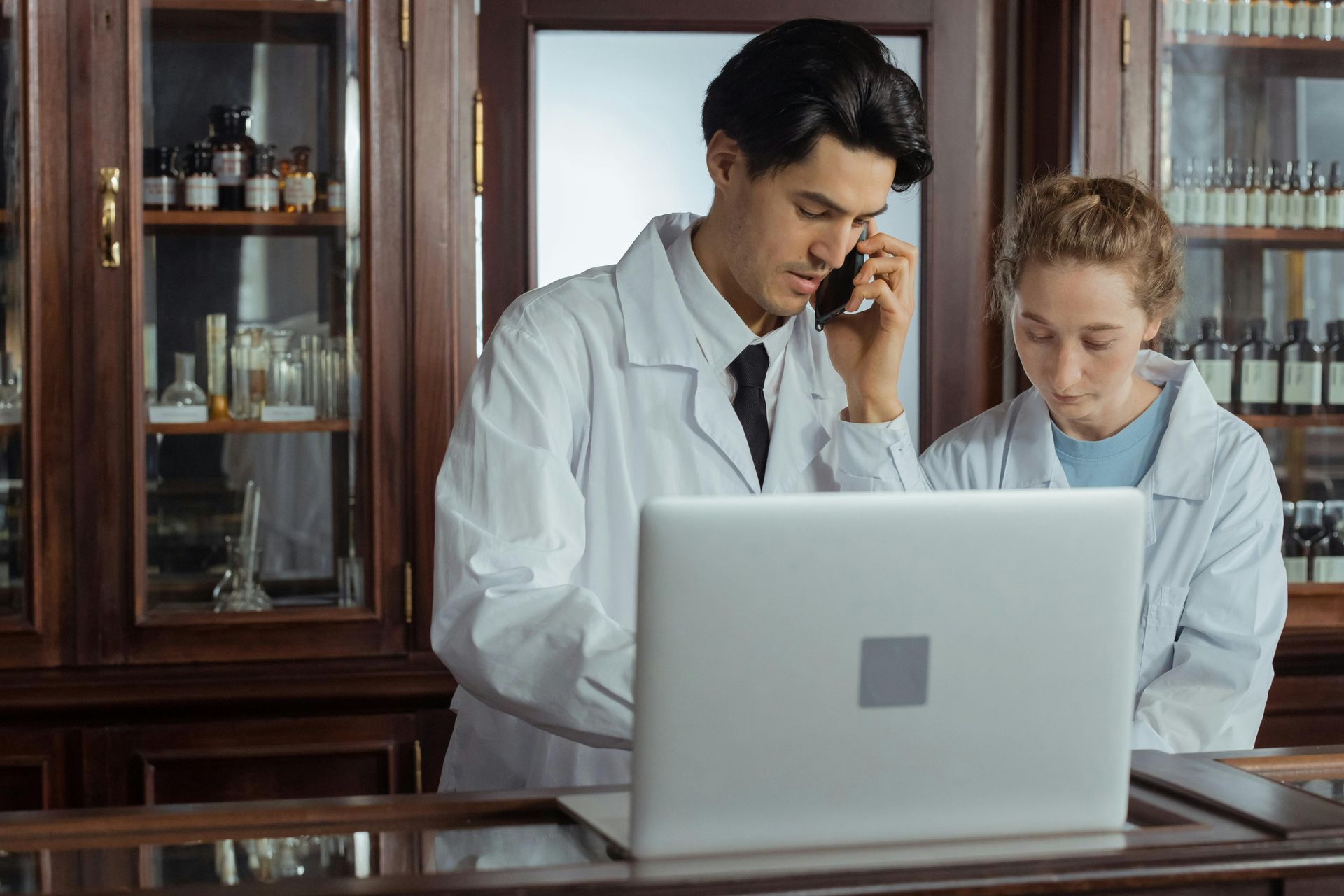 Two people in lab coats reviewing a laptop in a wood-paneled office, one speaking on a phone.