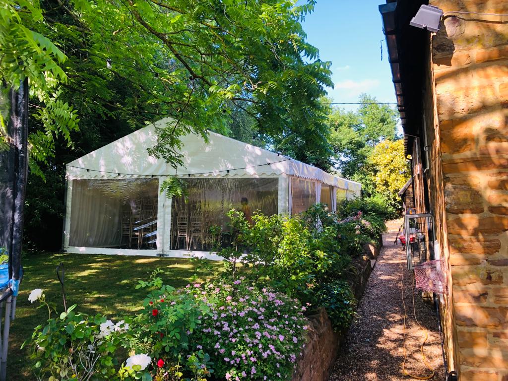 A large white tent is sitting in the middle of a garden next to a brick building.
