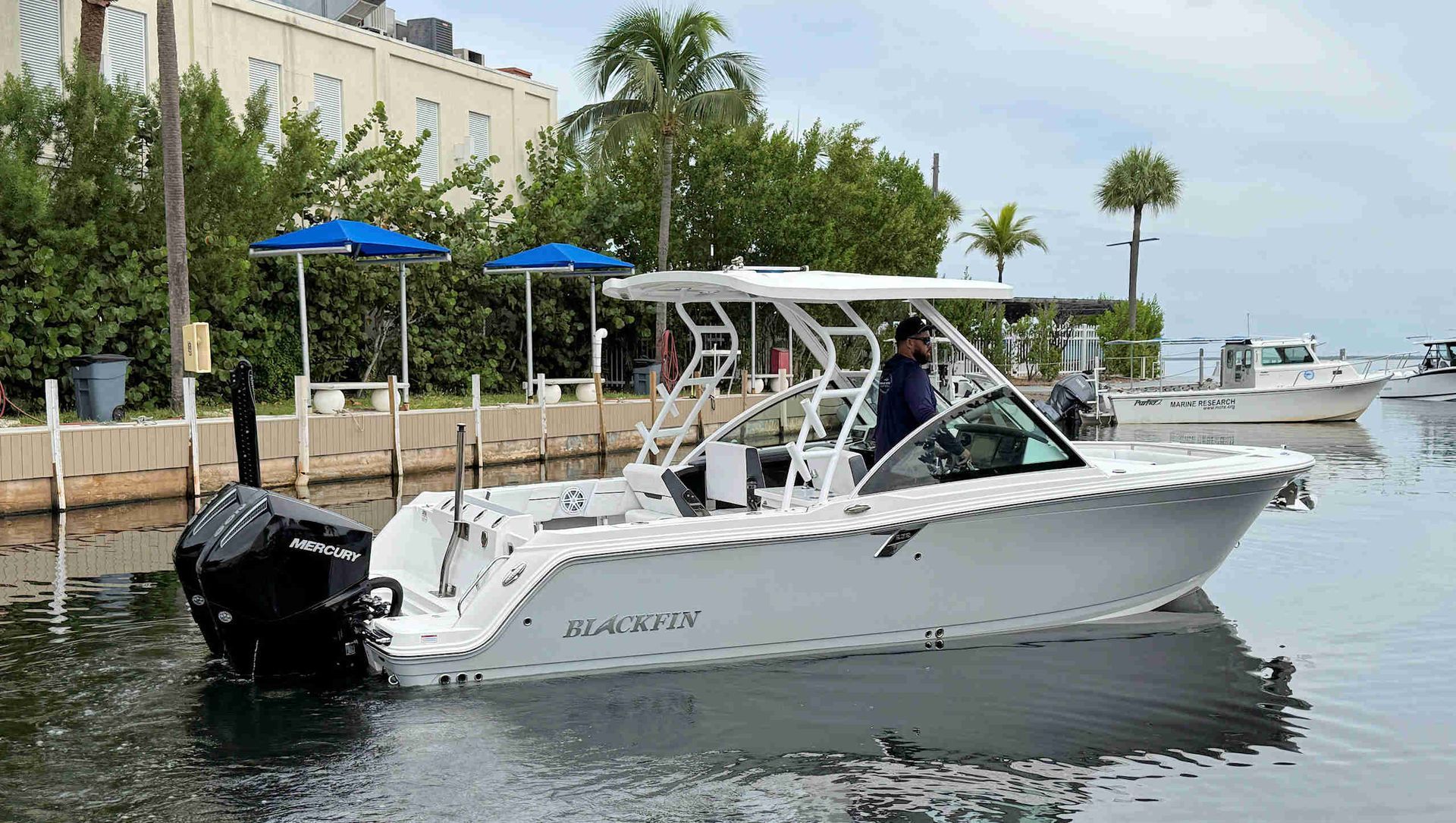 White motorboat docked on calm water beside a waterfront building with palm trees and blue umbrellas.