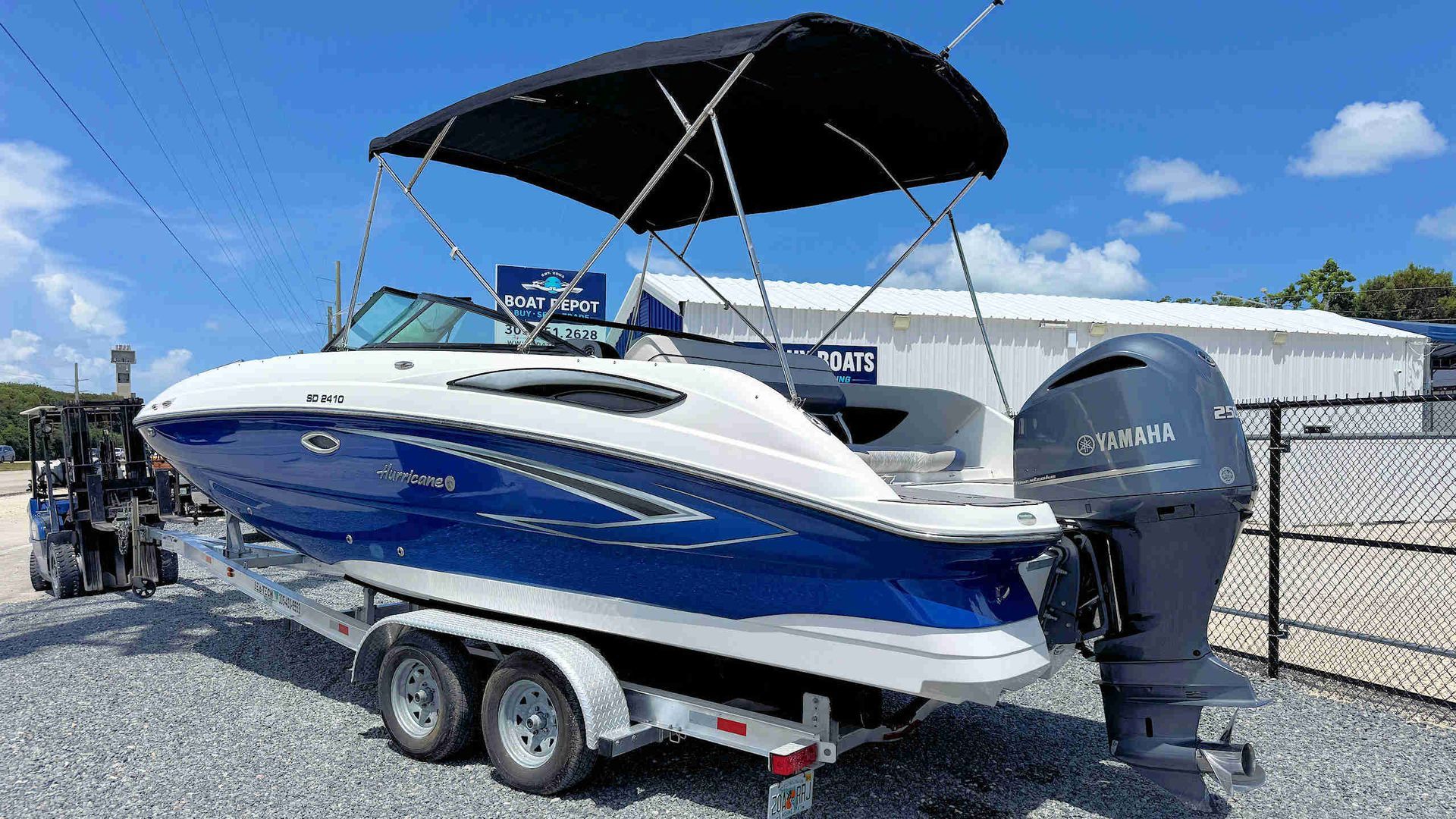 White and blue motorboat on a trailer with a black canopy, parked outdoors on gravel under a blue sky.