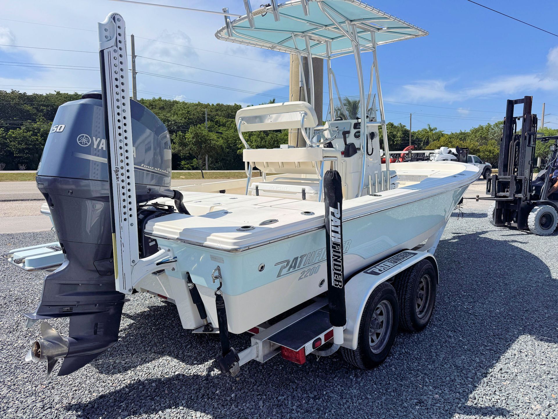 White fishing boat on a trailer with outboard motor, parked on gravel near a boatyard lift.