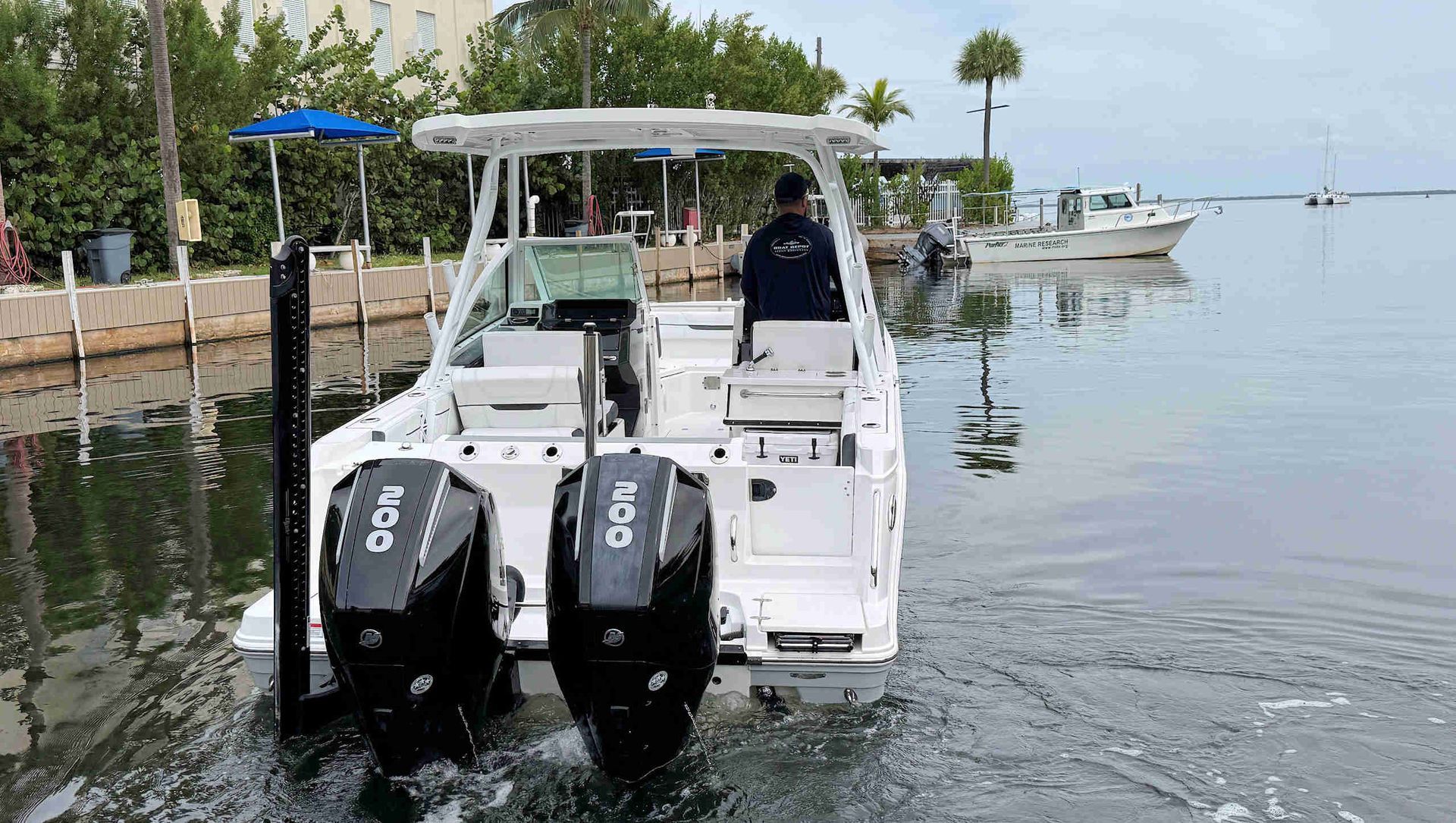 Small white motorboat with twin outboards in a calm canal, heading away from a dock on a cloudy day