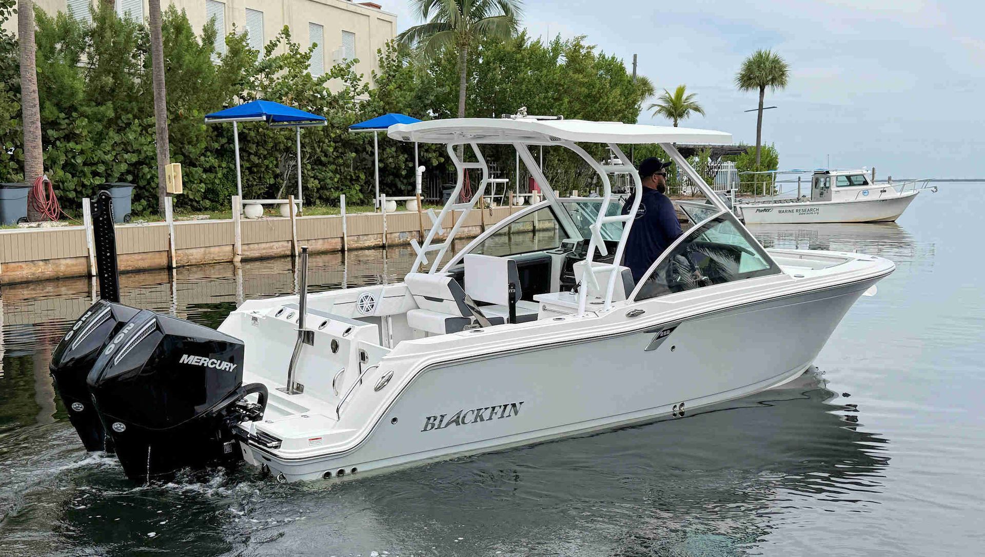 White dual-console fishing boat docked on calm water beside a marina, with a blue canopy and black outboard motor