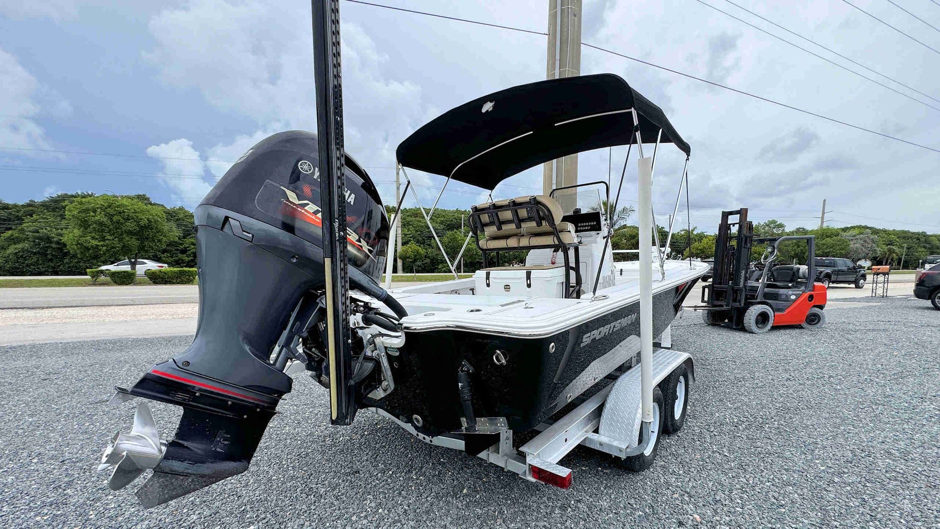 Black fishing boat on gravel with black bimini top and outboard motor, near a forklift under cloudy sky