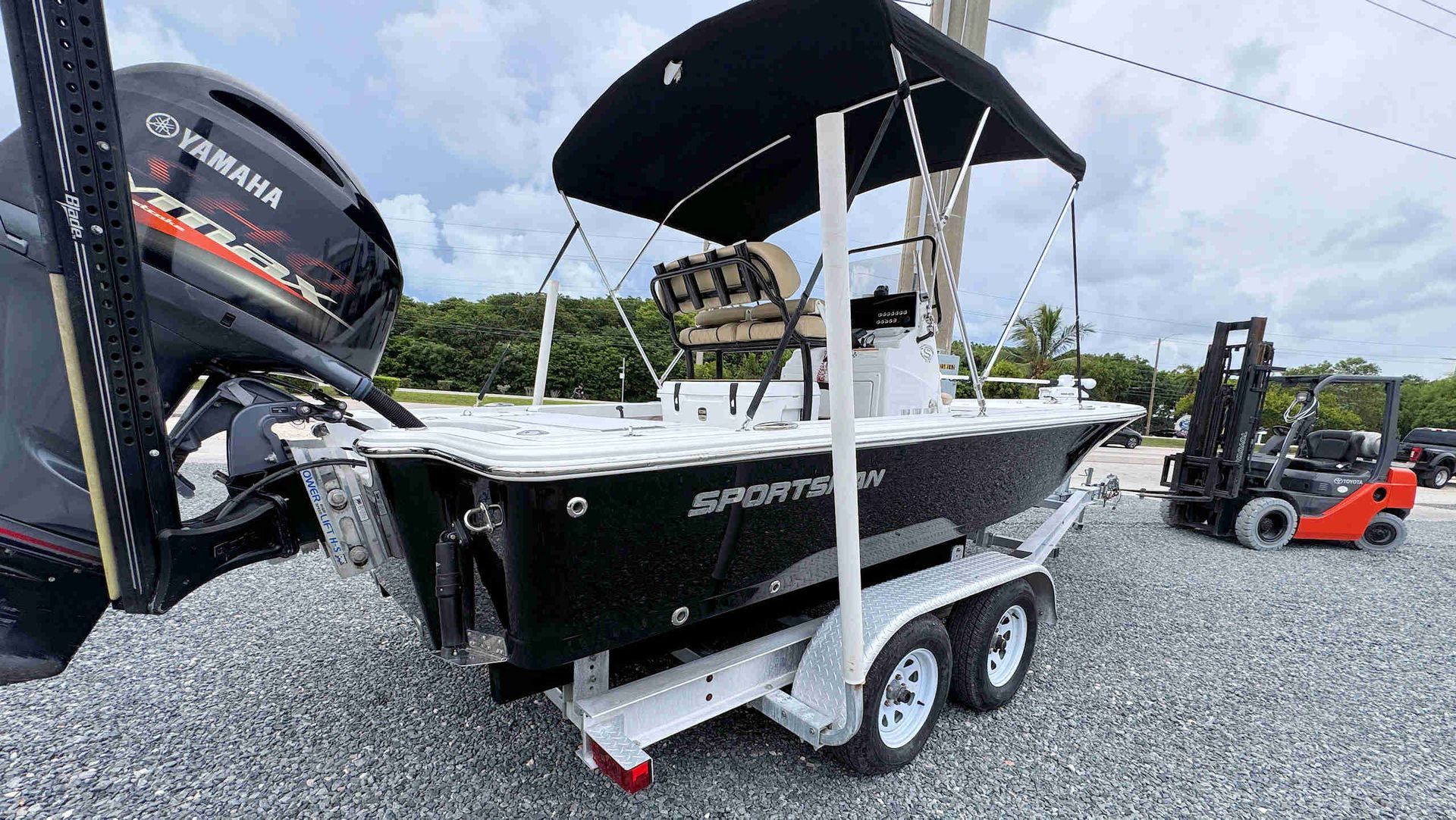 Black fishing boat on a trailer beside a large black outboard motor, parked on gravel near water.