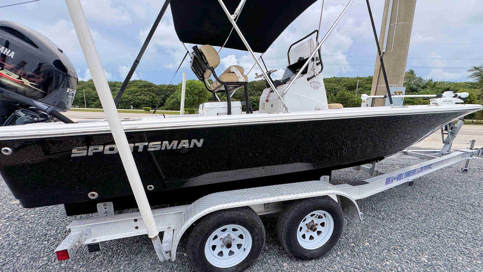 Black Sportsman boat on trailer at a gravel lot, with bimini top and outboard motor under a cloudy sky