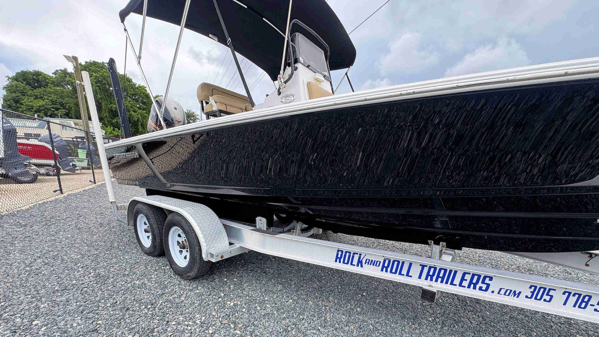 Black boat on an aluminum tandem axle trailer in a gravel lot under a canopy