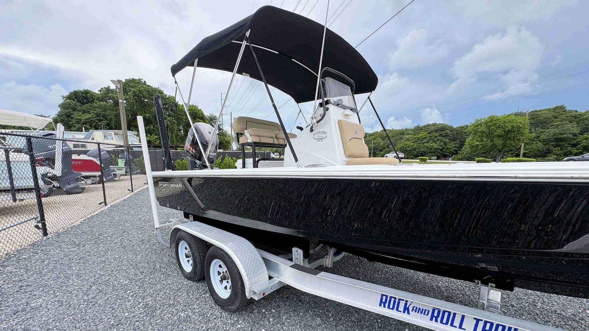 Black boat on a trailer with a black canopy, parked on gravel beside a fenced lot under a cloudy sky