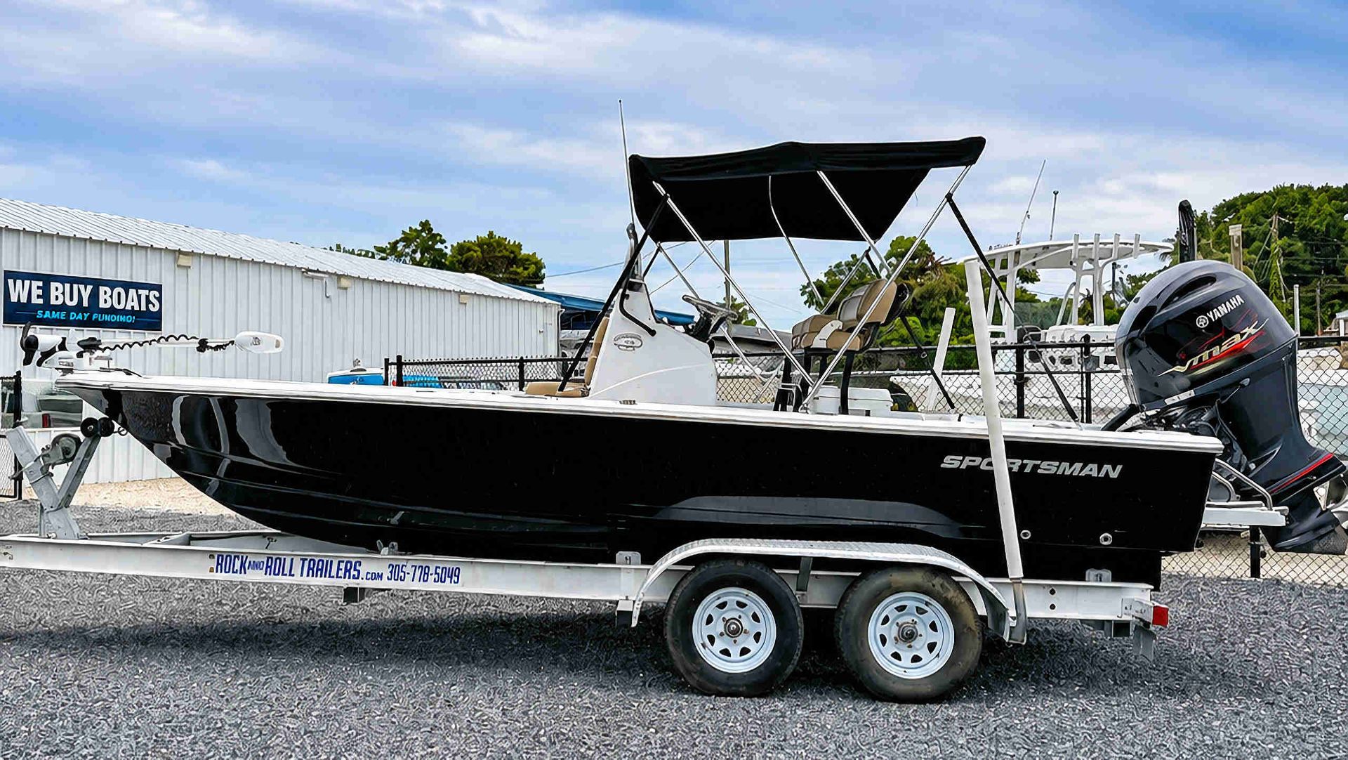 Black center-console fishing boat on a trailer in a gravel lot, with outboard motor and canopy.