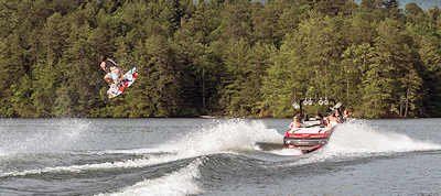 A person does a flip while wakeboarding behind a boat on a lake with a treeline in the background.