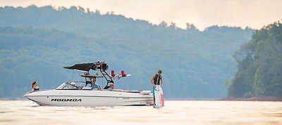 A white speedboat with people on board, wakeboarding on a lake, with a hilly landscape in the background.